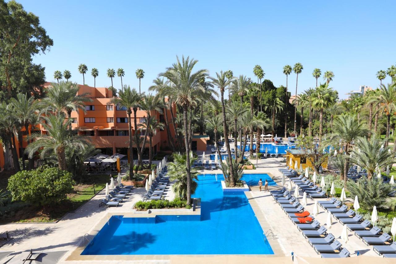 Kenzi Rose Garden Hotel Marrakesh Exterior photo The photo shows a resort-style area with several swimming pools surrounded by palm trees. The pools are clear blue and are lined with numerous lounge chairs for relaxation. There is a vibrant atmosphere, highlighted by lush greenery and a sunny sky. 