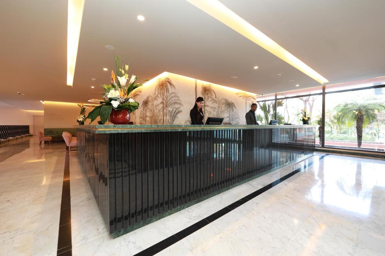 Kenzi Rose Garden Hotel Marrakesh Exterior photo The image shows a modern reception area, likely for a hotel or office building. There is a stylish front desk made of dark materials with a smooth surface. Behind the desk, two staff members are engaged in their work, possibly checking in guests or a